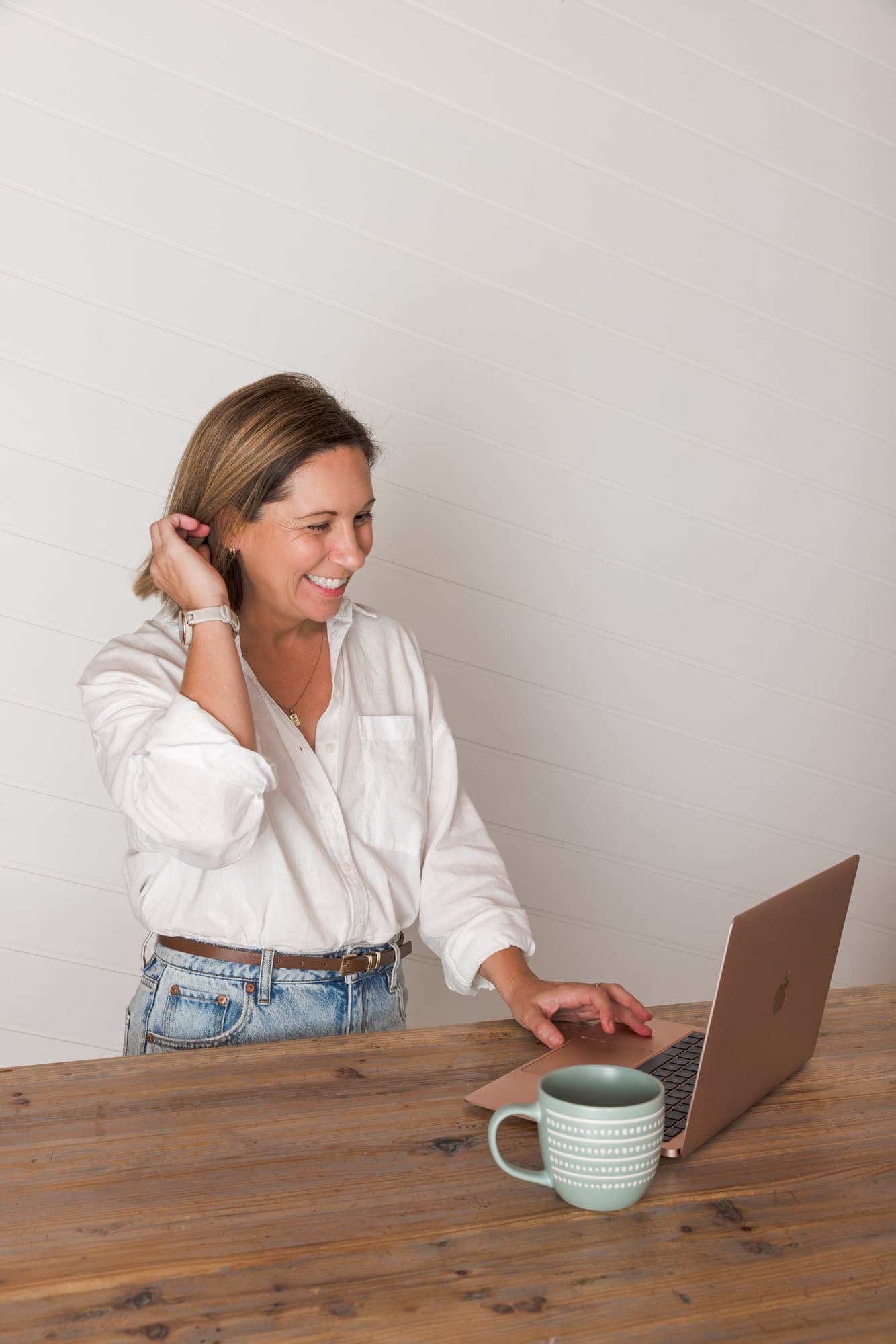Steph standing at her timber desk, one hand on the laptop trackpad, laughing at the screen