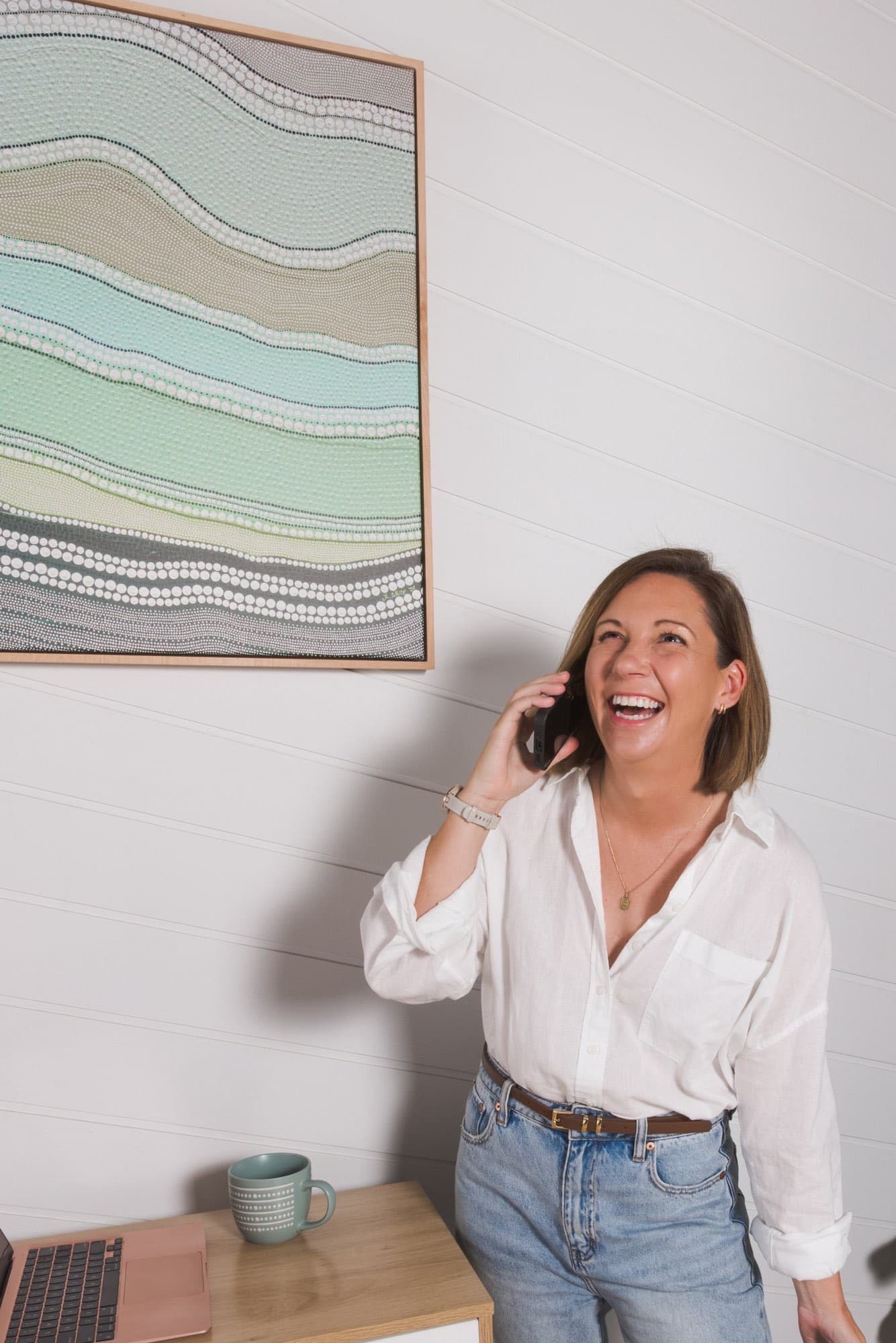 Steph mid-laugh on a phone call beside her desk, the dot-art painting on the wall behind her